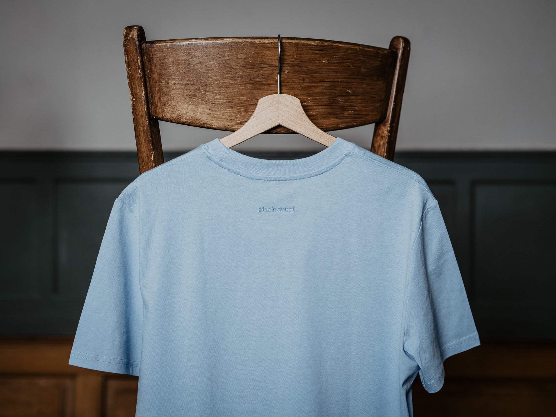 T-Shirt in blau hängt am Stuhl und zeigt stich.wort Logo auf der Rückseite