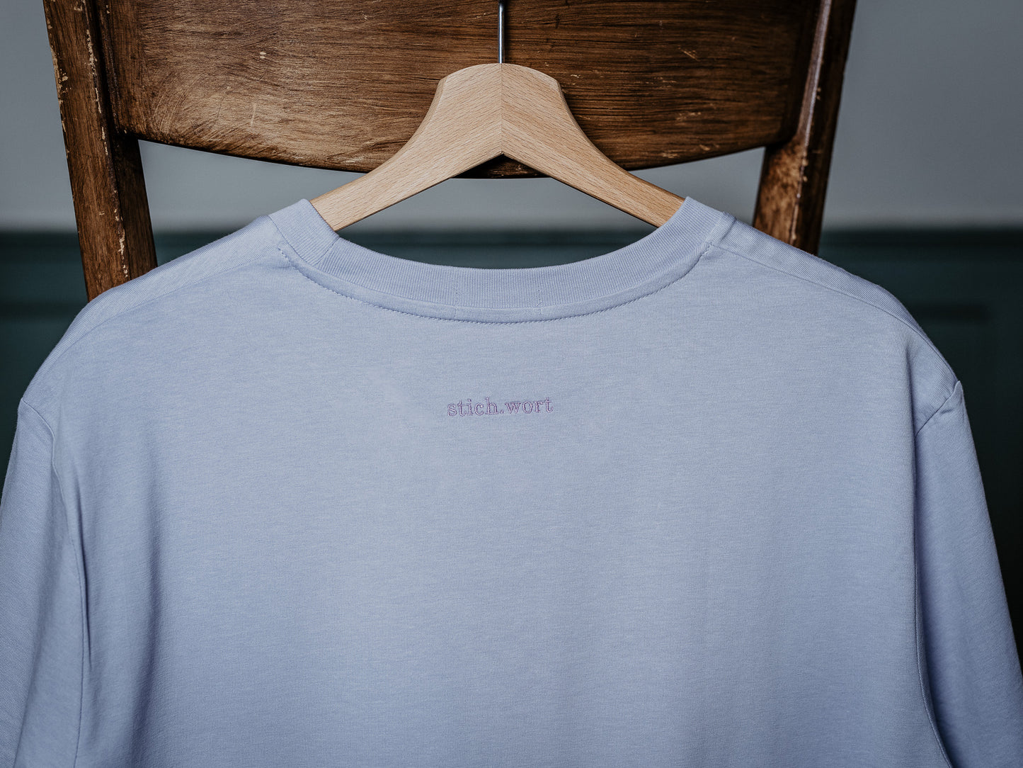 T-Shirt in liila hängt am Stuhl und zeigt stich.wort Logo auf der Rückseite