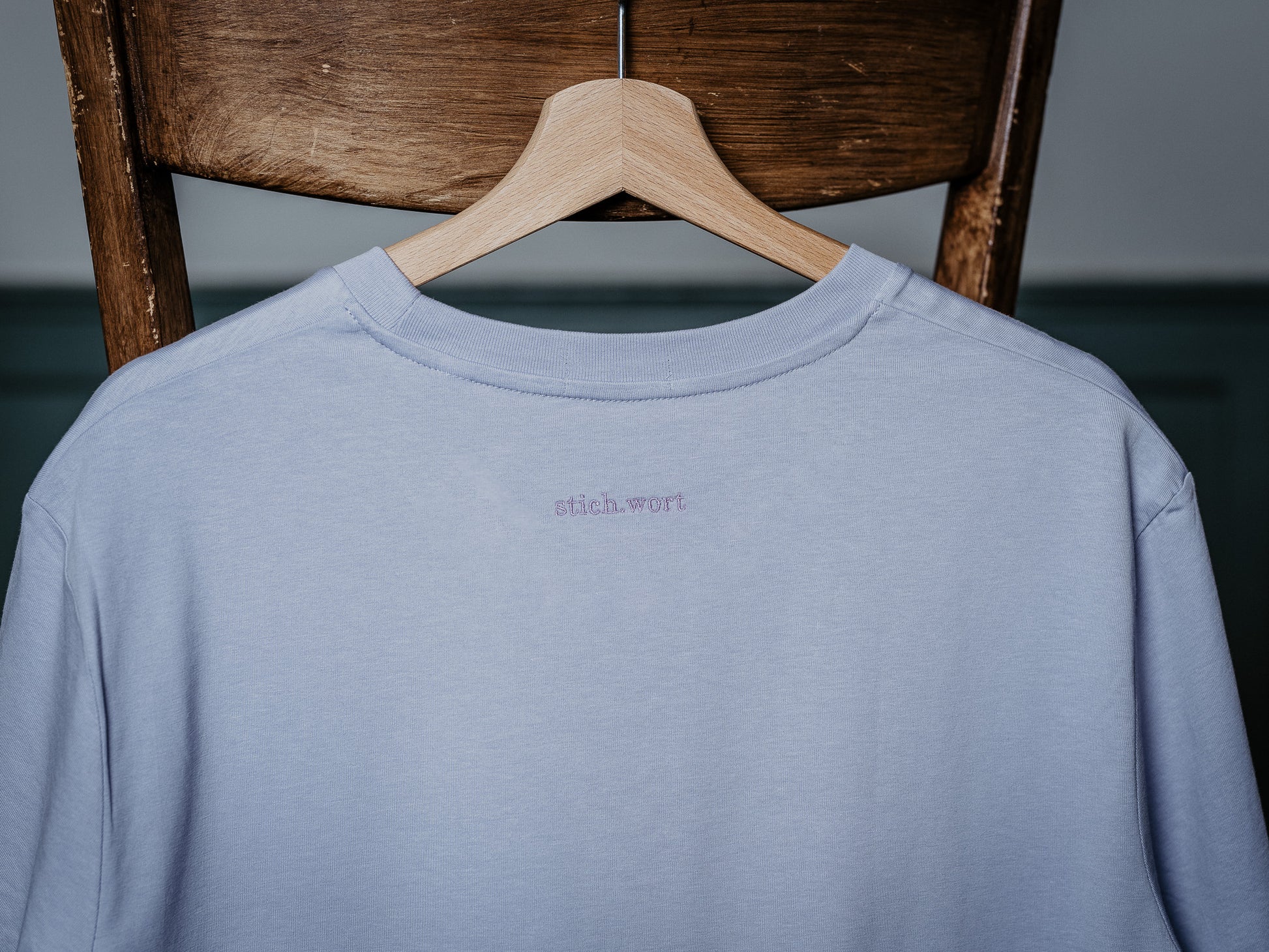 T-Shirt in liila hängt am Stuhl und zeigt stich.wort Logo auf der Rückseite